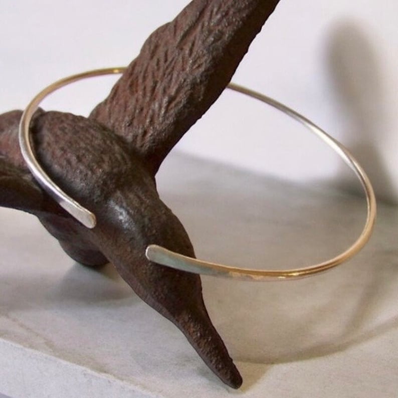 May include: A silver-colored open cuff bracelet. The bracelet is a simple, minimalist design with a smooth, polished finish. The ends of the bracelet are slightly flattened and open. The bracelet is displayed on a dark brown, textured surface.
