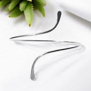 May include: A silver, open-ended, twisted bangle bracelet. The bracelet is made of a single, thin piece of metal that is twisted into a spiral shape.