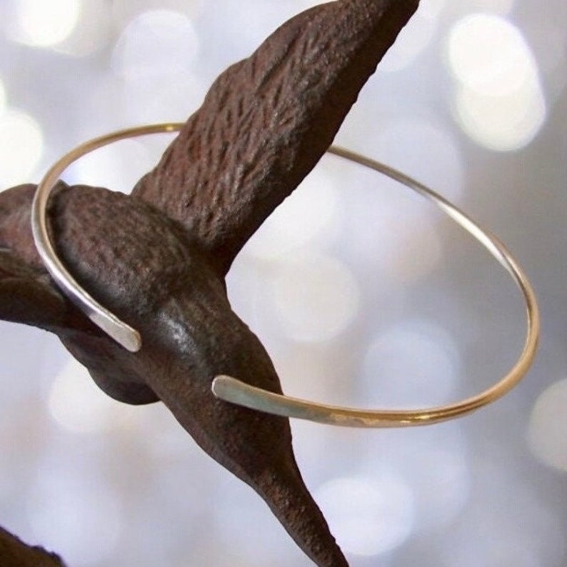 May include: A delicate, open-ended silver-toned bracelet. The bracelet has a simple, minimalist design with a smooth, polished finish. The bracelet is displayed on a dark, textured surface, creating a contrast with the bright metal.