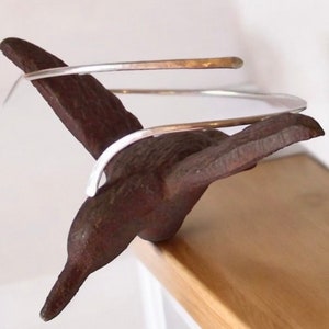 May include: A silver wire choker necklace with a hammered finish. The necklace is displayed on a brown metal hummingbird figurine.