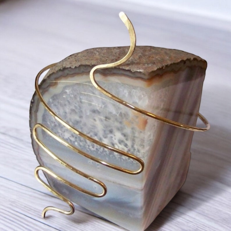 Pu&ograve; includere: Un braccialetto in metallo dorato con un design a spirale. Il braccialetto &egrave; esposto su una pietra di agata marrone e bianca.