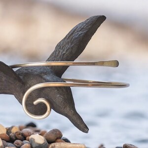 May include: A gold-toned metal bangle bracelet with a simple, curved design. The bracelet is shown on a dark metal bird sculpture.