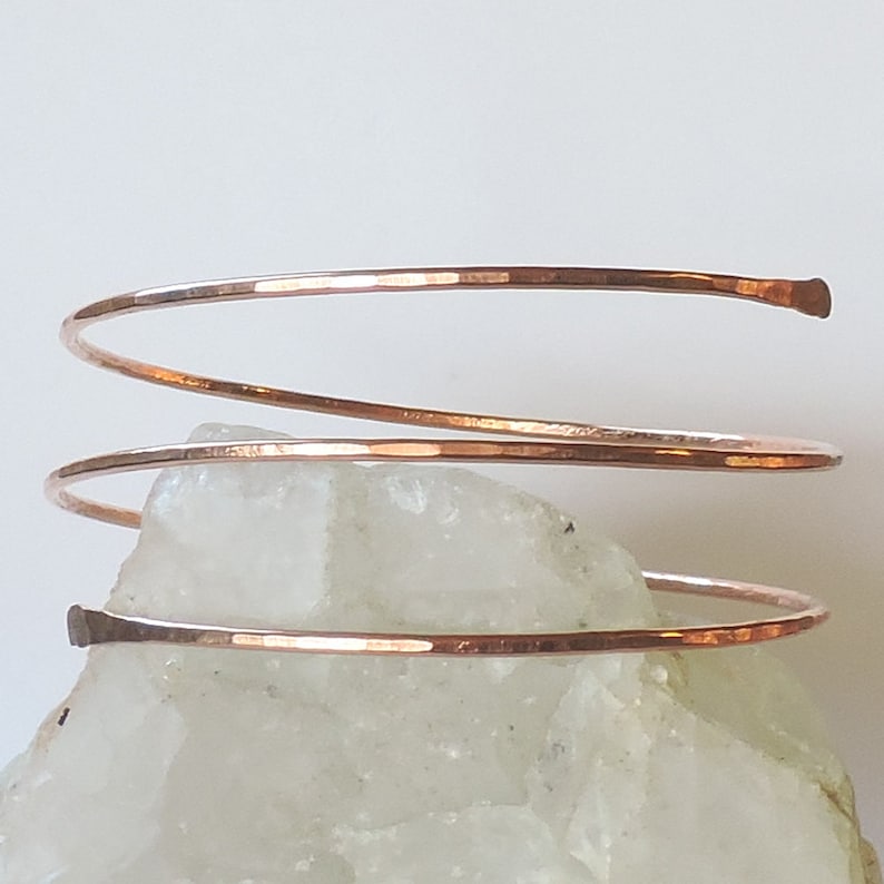 May include: A hammered copper bangle bracelet with a spiral design. The bracelet is made of a single piece of wire that is wrapped around itself to create the spiral effect.