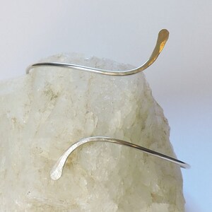 May include: A silver-toned metal cuff bracelet with a thin, curved wire design. The bracelet features two ends that taper into flattened, slightly hammered tips, creating an open, asymmetrical silhouette that wraps around the wrist area.