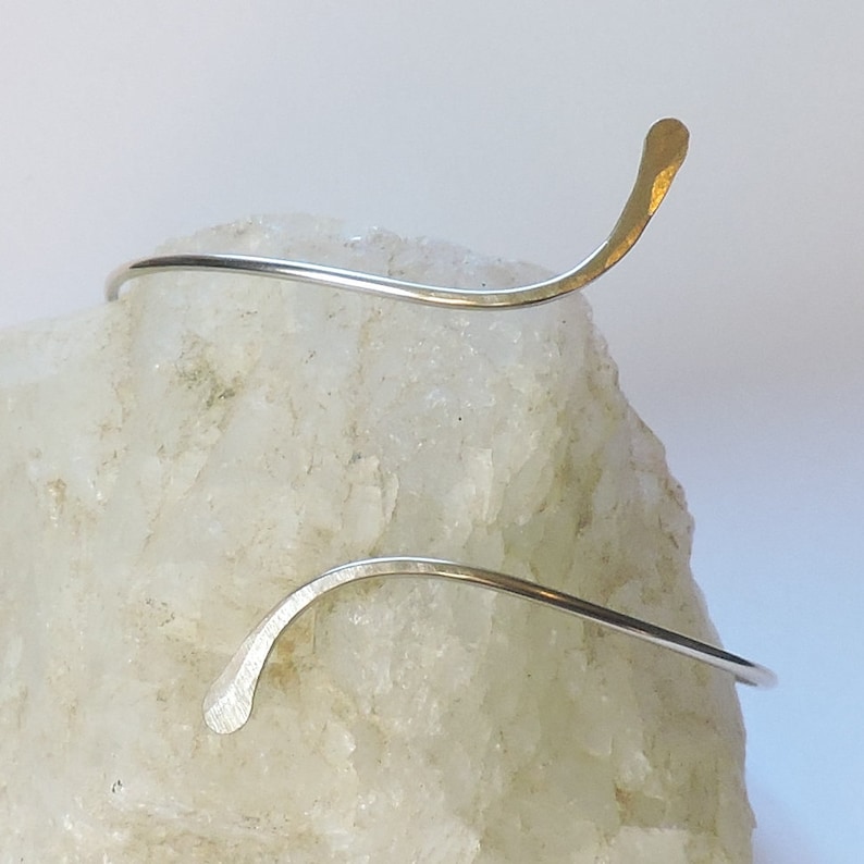 May include: A silver-toned metal cuff bracelet with a thin, curved wire design. The bracelet features two ends that taper into flattened, slightly hammered tips, creating an open, asymmetrical silhouette that wraps around the wrist area.