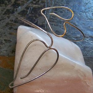 May include: A silver-toned metal hair accessory featuring a delicate design of two heart-shaped elements. The accessory is positioned on a light pink, textured surface, creating a contrast in color and texture. The design is simple yet elegant.