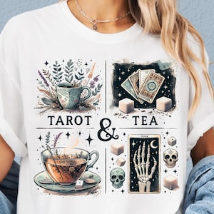 May include: White t-shirt with a graphic design featuring the words "TAROT & TEA." The design includes illustrations of teacups, tarot cards, sugar cubes, a skeleton hand, and skulls. The overall theme is mystical and whimsical.