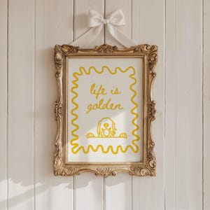May include: A gold framed print with a white background and yellow text that reads "life is golden". A yellow outline of a dog's head is peeking over the top of the text.