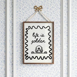 May include: A rectangular wall art print in a brown wooden frame, featuring a black wavy border on a white background. The text reads "life is golden" above a line drawing of a dog peeking over a ledge. A cream ribbon and gold bow hang it.