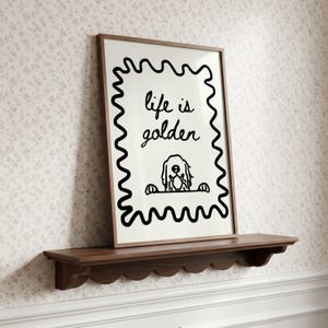 May include: A framed wall art print featuring a black wavy border on a white background. The text reads life is golden in a handwritten script font. Below the text is a simple black line drawing of a dog peeking over a horizontal line.