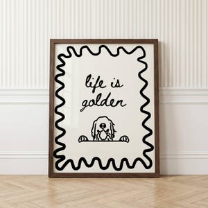 May include: A framed wall art print featuring a cream-colored background with a thick black wavy border. The text "life is golden" is written in black cursive script above a simple black line drawing of a dog peeking over a horizontal line.