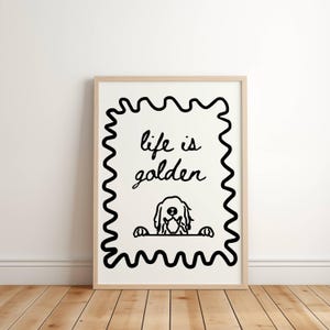 May include: A framed wall art print featuring a black line drawing on a white background. The design includes a wavy rectangular border, the text "life is golden" in a cursive script, and a stylized illustration of a dog peeking over a line.