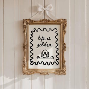 May include: A rectangular gold ornate frame containing a white print with a black wavy border. The text reads "life is golden" above a black line drawing of a dog peeking over a ledge. A white satin ribbon bow is attached to the top of the frame.