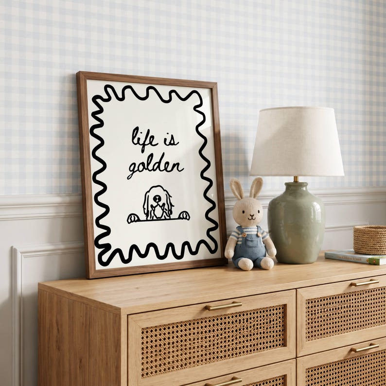 May include: A framed wall art print with a dark wood frame featuring a black wavy border on a cream background. The text reads "life is golden" above a black line drawing of a dog peeking over a ledge. The print is displayed on a wooden dresser.
