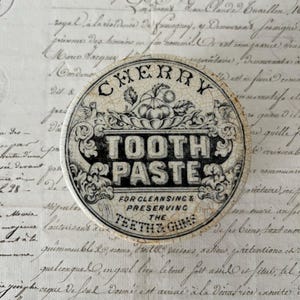 May include: A round, cream-colored ceramic lid with a crackled finish, featuring black printed text that reads CHERRY TOOTH PASTE FOR CLEANSING & PRESERVING THE TEETH & GUMS, decorated with a central illustration of cherries and ornate scrollwork.