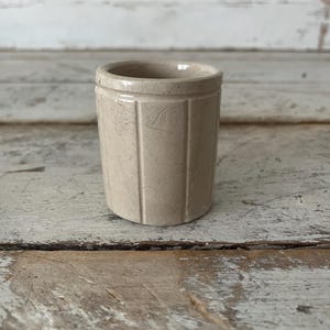 May include: A small, cylindrical beige ceramic vessel featuring a series of vertical recessed grooves along its exterior surface. The item has a smooth, glazed finish with subtle surface texture and a simple, open top rim design.