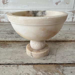 May include: A footed bowl crafted from polished cream-colored stone with natural grey veining. The bowl features a wide, circular rim with three horizontal engraved lines and rests on a spherical pedestal base atop a circular, tiered foot.