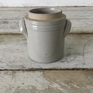 May include: A light gray stoneware crock featuring a cylindrical body, two small side handles, and a raised rim with a smooth, unglazed beige top edge. The vessel has a simple, functional design with a uniform matte finish and a sturdy base.