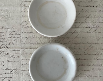 May include: Two small, round, white ceramic dishes with smooth, slightly raised edges. Both dishes feature a simple, minimalist design with a glossy finish and subtle, light brown circular staining visible on the interior base of each piece.