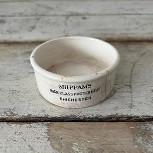 May include: A small, off-white ceramic bowl with a slightly aged appearance. The bowl features the text "SHIPPAM'S HIGH CLASS POTTED MEAT CHICHESTER" in black lettering. The bowl sits on a weathered, light-colored wooden surface.