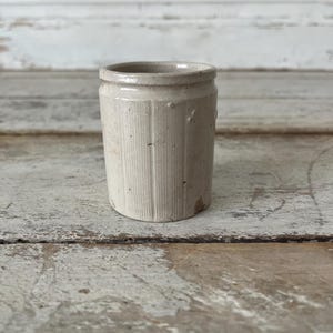 May include: A small, cylindrical, off-white ceramic crock featuring a textured, vertically ribbed exterior surface. The vessel has a simple, slightly flared rim at the top and shows signs of wear with minor surface imperfections and discoloration.