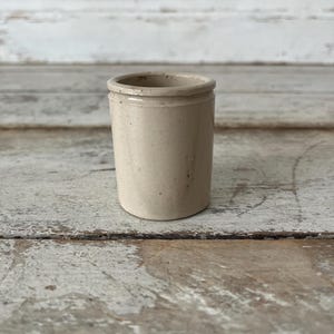 May include: A small, cylindrical, beige stoneware pot with a smooth finish and a slightly raised rim at the top. The vessel features a simple, unadorned design with subtle variations in the light tan color across its exterior surface.