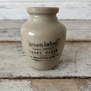 May include: A small, beige stoneware jar with a narrow neck and a wide rim. The front features black printed text that reads: green label Genuine INDIAN CURRY PASTE J.A. SHARWOOD & Co. Ltd LONDON. The surface has a smooth, glazed finish.