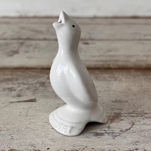 May include: A white ceramic bird-shaped pie vent with an open beak and a small black eye. The base of the figure features the embossed text LE CREUSET. The bird has a smooth, glossy finish and is designed to stand upright on a flat surface.