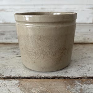 May include: A cylindrical stoneware crock featuring a light beige glaze with a visible network of fine cracks across the surface. The vessel has a smooth, slightly flared rim and a thick, sturdy base, presenting a weathered and aged appearance.