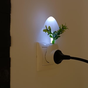 Könnte beinhalten: Ein weißes, pilzförmiges LED-Nachtlicht, das in eine Steckdose eingesteckt ist. Die Leuchte hat einen durchscheinenden weißen Schirm, der ein sanftes Licht abgibt, und ist mit kleinen grünen Kunstblättern an der Basis verziert. Ein schwarzes Kabel.