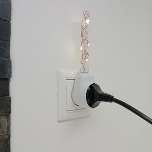 Könnte beinhalten: Ein weißer Wandsteckdosenadapter mit einem durchsichtigen Glasröhrchen, das mit dünnem Kupferdraht und kleinen leuchtenden LED-Lichtern gefüllt ist. Ein schwarzer Netzstecker steckt seitlich im Adapter, der in der Wand eingesteckt ist.