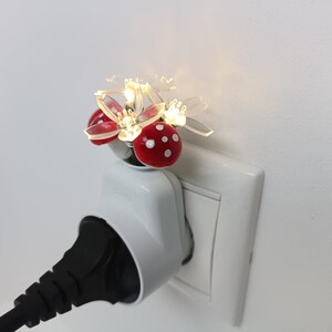 Op de afbeelding: Een nachtlampje voor in het stopcontact met een cluster van doorzichtige, bloemvormige blaadjes en kleine rode paddenstoelhoedjes met witte stippen. Het lampje brandt en geeft een warm schijnsel terwijl het in een wit stopcontact zit.