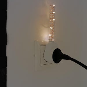 May include: A white plug-in night light featuring a clear cylindrical glass tube with an internal copper-colored spiral filament that emits a warm glow, inserted into a wall outlet alongside a black power cord and plug.
