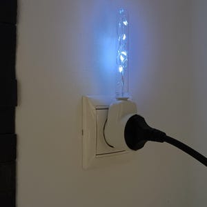 May include: A white wall-mounted plug-in night light featuring a clear, cylindrical glass tube filled with glowing cool white LED fairy lights. The device is inserted into a standard wall outlet, with a black power cord plugged into its front.