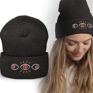 Puede incluir: Gorro gris oscuro con un diseño bordado de tres ojos rojos con rayos amarillos. El gorro está hecho de un material suave y tejido. El diseño está centrado en el borde doblado.