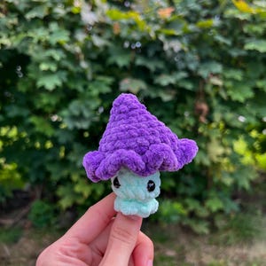 May include: A small crocheted mushroom toy with a purple cap and a light blue body. The mushroom has black eyes and a small mouth.