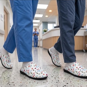 Peut inclure: Sabots blancs à enfiler avec un motif médical coloré composé de stéthoscopes, seringues, cœurs et pilules. Les chaussures ont une semelle noire et sont portées avec des chaussettes blanches et des pantalons de bloc bleu dans un couloir.