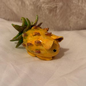 May include: A small, yellow, handmade pineapple figurine with green leaves and a black eye. The pineapple has a textured surface and is made of clay.