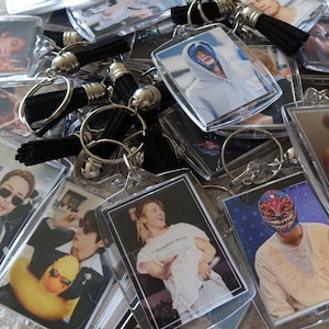 Puede incluir: Una colección de llaveros rectangulares de acrílico con diversas impresiones fotográficas de personas, incluida una que lleva una máscara colorida. Cada llavero está unido a una anilla de metal plateado y una borla de tela negra corta.