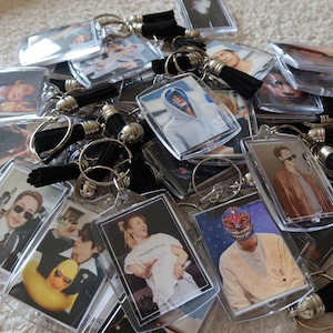 Puede incluir: Una colección de llaveros rectangulares de acrílico con diversas impresiones fotográficas de personas. Cada llavero está unido a una anilla de metal plateado e incluye una borla corta de tela negra que cuelga del punto de sujeción superior.