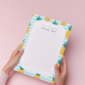 May include: A lined notepad with a pineapple and blue striped border. The notepad has the text "FROM THE DESK OF Marioly Said" at the top. The notepad is held by two hands against a pink background.