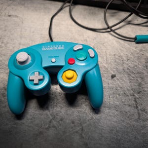 May include: A teal Nintendo GameCube controller with a white analog stick, a yellow button, and red, green, and gray buttons. The controller has a cross-shaped directional pad and a cable with a teal connector.