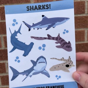 May include: A sheet of stickers featuring various shark illustrations. The stickers are on a white background with blue borders. The text "SHARKS!" is at the top of the sheet and "FUNKY FROG FRANCHISE" is at the bottom.