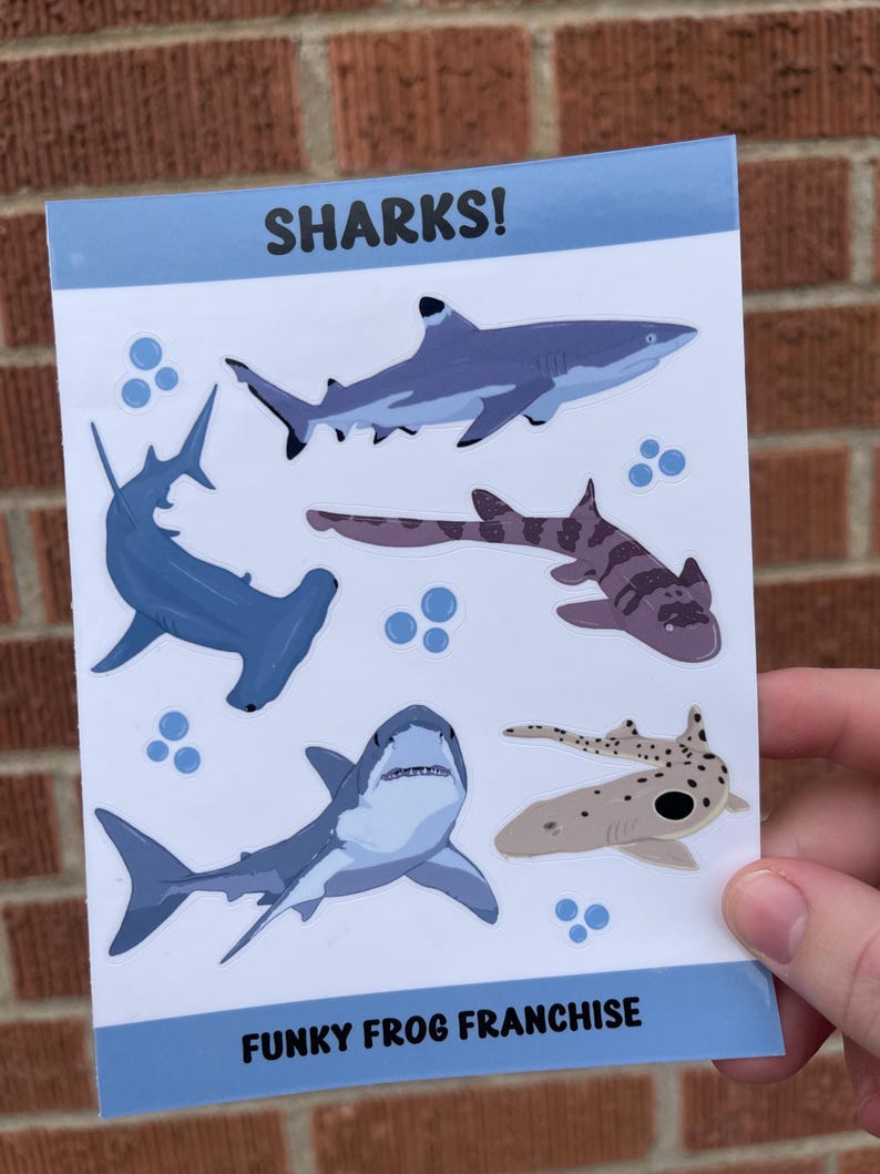 May include: A sheet of stickers featuring various shark illustrations. The stickers are on a white background with blue borders. The text "SHARKS!" is at the top of the sheet and "FUNKY FROG FRANCHISE" is at the bottom.
