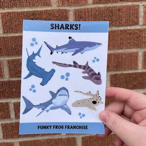 May include: A sheet of stickers featuring various cartoon sharks in blue, gray, and brown colors. The stickers are on a white background with blue dots. The sheet has the text "SHARKS!" at the top and "FUNKY FROG FRANCHISE" at the bottom.