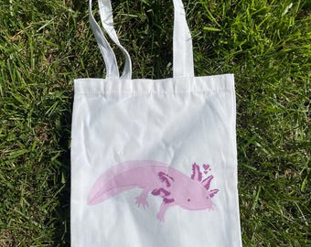 May include: White tote bag featuring a pink axolotl illustration. The axolotl is shown in profile with detailed gills and a heart design. The bag has two white handles and is placed on green grass.