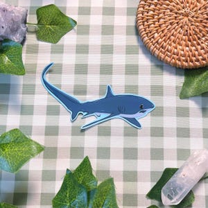 May include: A blue shark sticker with a light blue outline. The sticker is on a green and white checkered surface, surrounded by green leaves, crystals, and a woven coaster.
