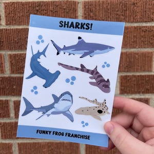 May include: A sheet of stickers featuring various shark illustrations. The stickers are on a white background with blue accents. The text "SHARKS!" is at the top of the sheet and "FUNKY FROG FRANCHISE" is at the bottom.