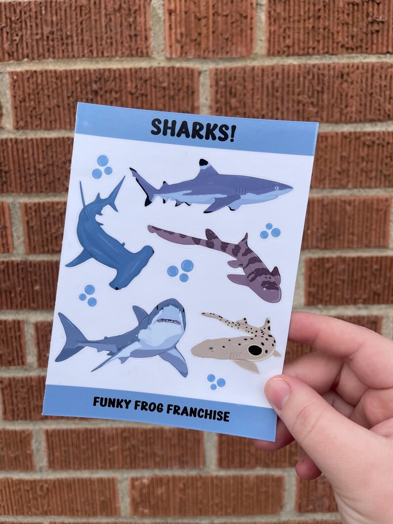 May include: A sheet of stickers featuring various shark illustrations. The stickers are on a white background with blue accents. The text "SHARKS!" is at the top of the sheet and "FUNKY FROG FRANCHISE" is at the bottom.