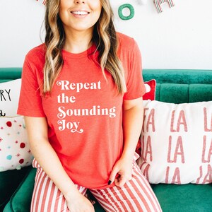 May include: A woman wearing a red t-shirt with the text "Repeat the Sounding Joy" in white. She is sitting on a green couch with pillows that say "Merry &" and "La La La".